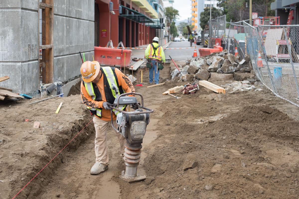 Construction worker operating a pneumatic compactor on excavated street soil during underground utility work.
