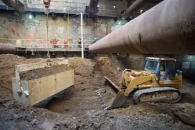 Construction equipment and excavation work inside underground transit station with temporary steel supports and large pipe.
