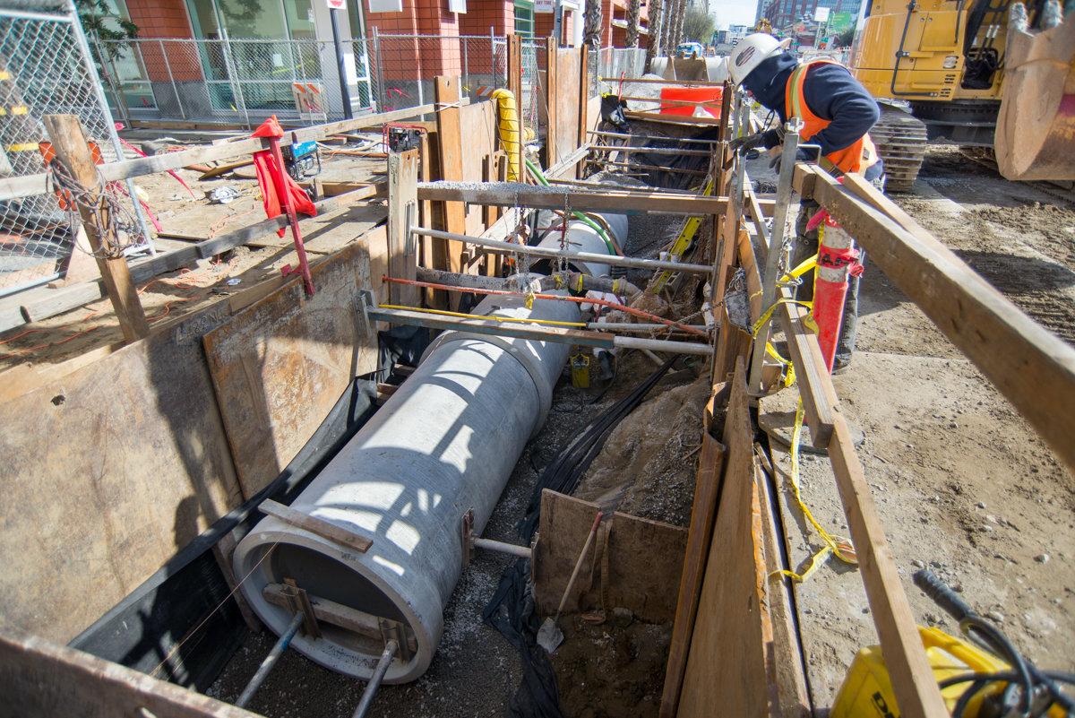 Workers install underground utility pipe in excavated trench on city street.