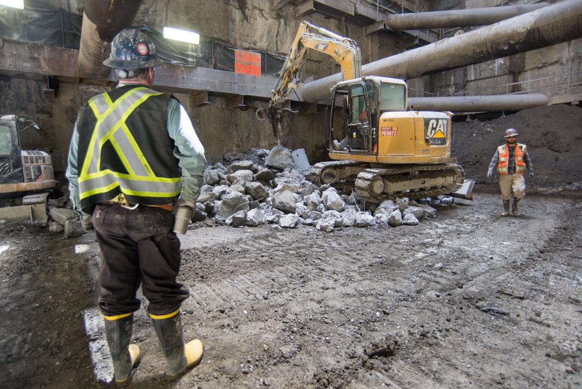 Workers in safety vests operate an excavator to break apart concrete tunnel sections at a sewer excavation site.