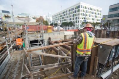 Worker in safety gear observing sheet pile wall and rebar installation at transit station construction site.