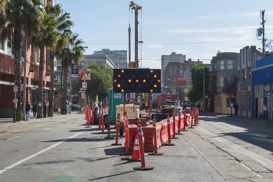 Construction zone with red and white barriers and directional signage on urban street surrounded by buildings.