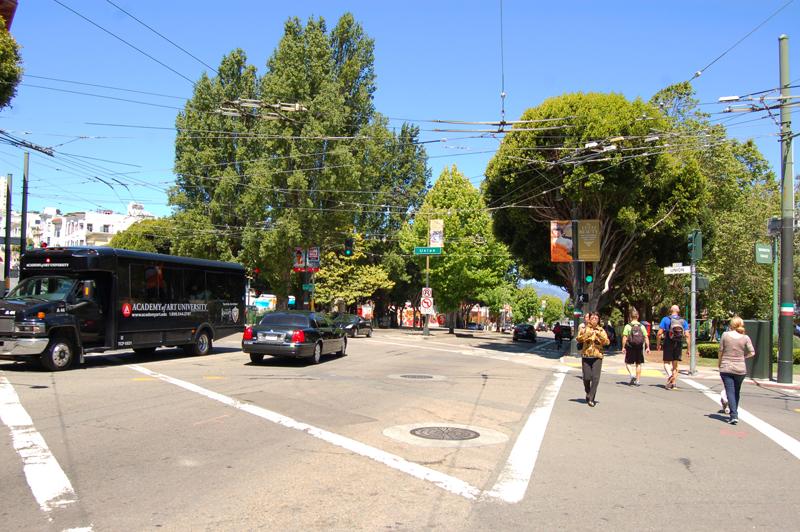 Busy street intersection with transit bus, pedestrians, and large trees under clear sky