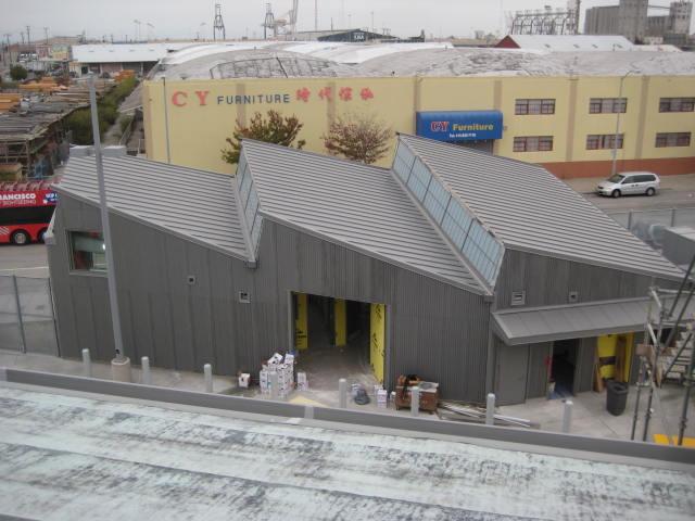 Gray industrial building with angled metal roof and yellow doors, waterfront location with cargo cranes visible in background.