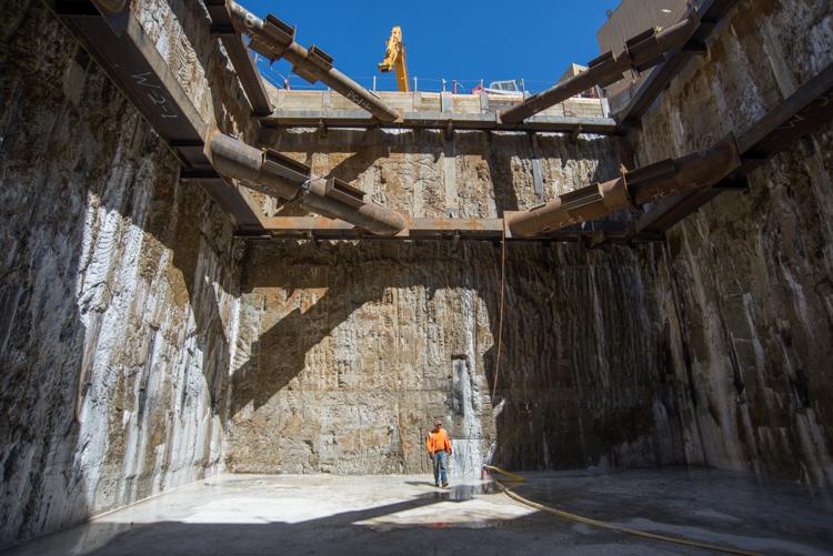 Worker in safety vest stands in large concrete underground shaft with steel beams overhead and construction equipment visible above.
