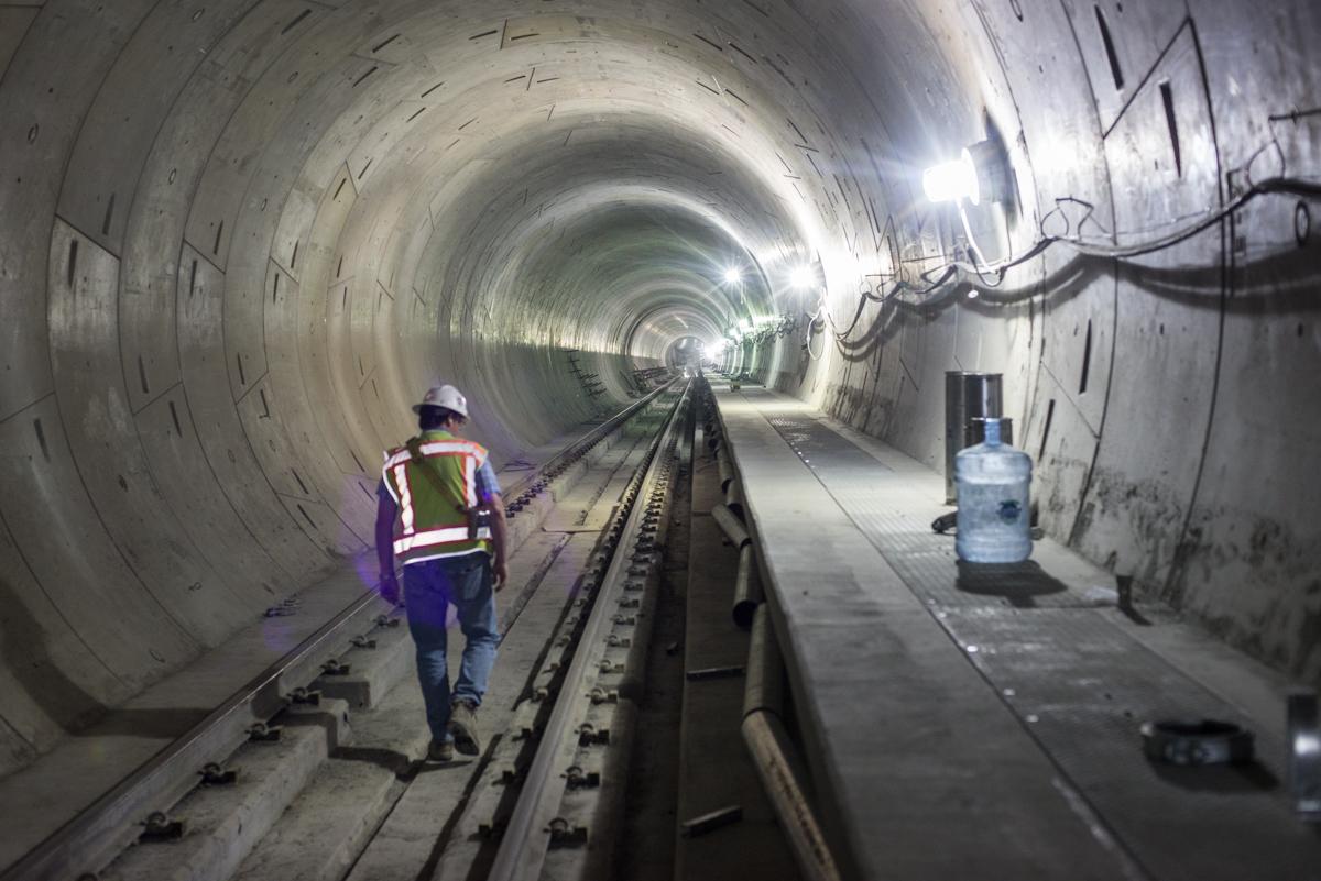 Worker in safety vest and hard hat walking along railroad tracks inside illuminated tunnel under construction.