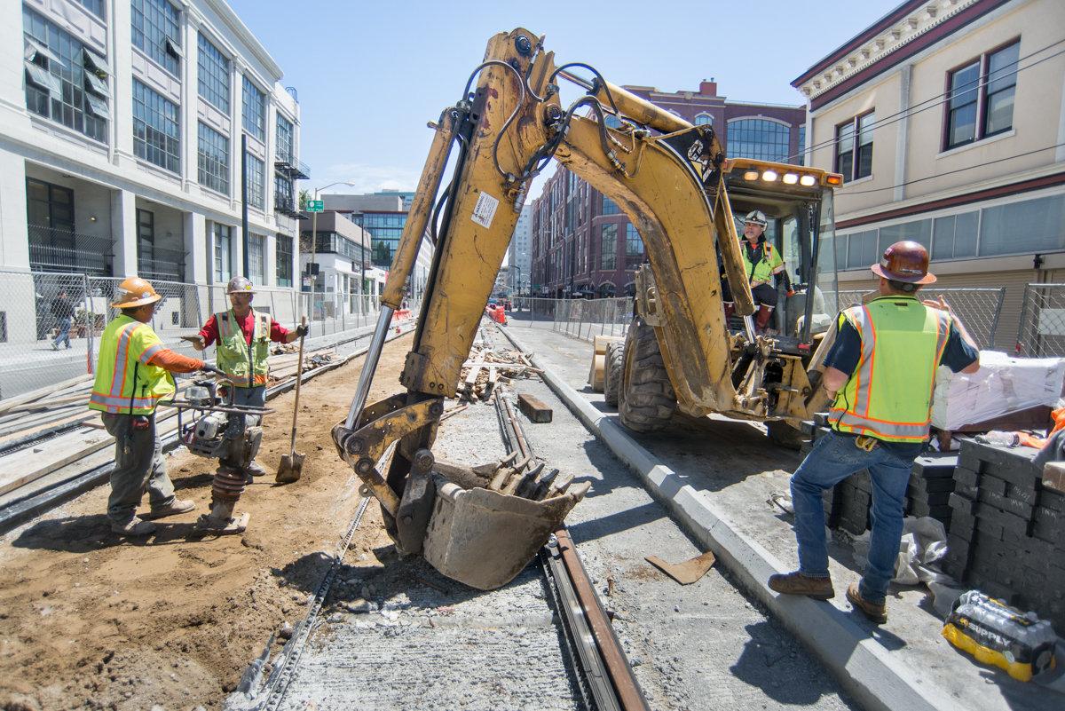 Construction crew operates excavator to backfill trench between dual surface trackways for Central Subway project.