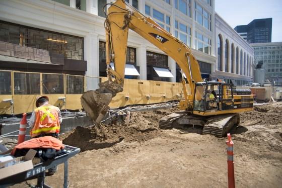 Construction excavator backfilling soil on urban street with workers and equipment present.