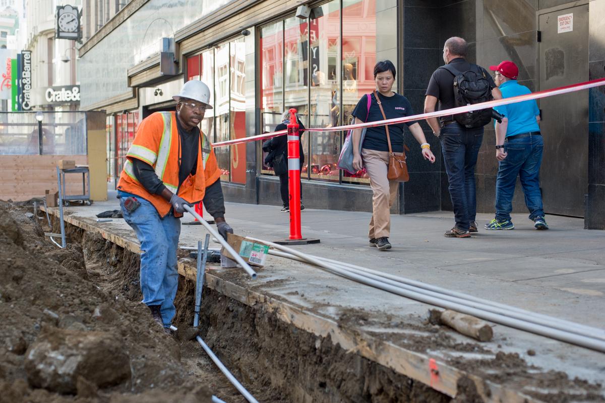 Construction worker in safety vest operates equipment in street trench while pedestrians pass behind barrier.