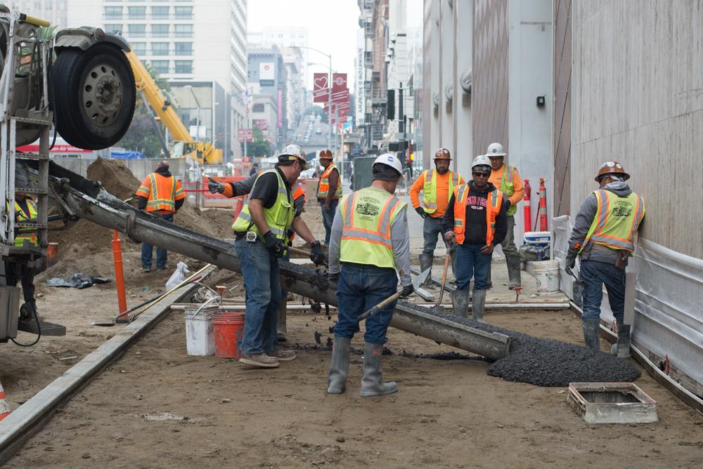 Construction workers in safety vests pour asphalt on Stockton Street sidewalk in downtown San Francisco.