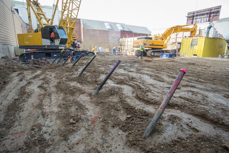 Construction site with tunnel boring machine, excavators, and purple pipes in muddy ground near brick buildings.