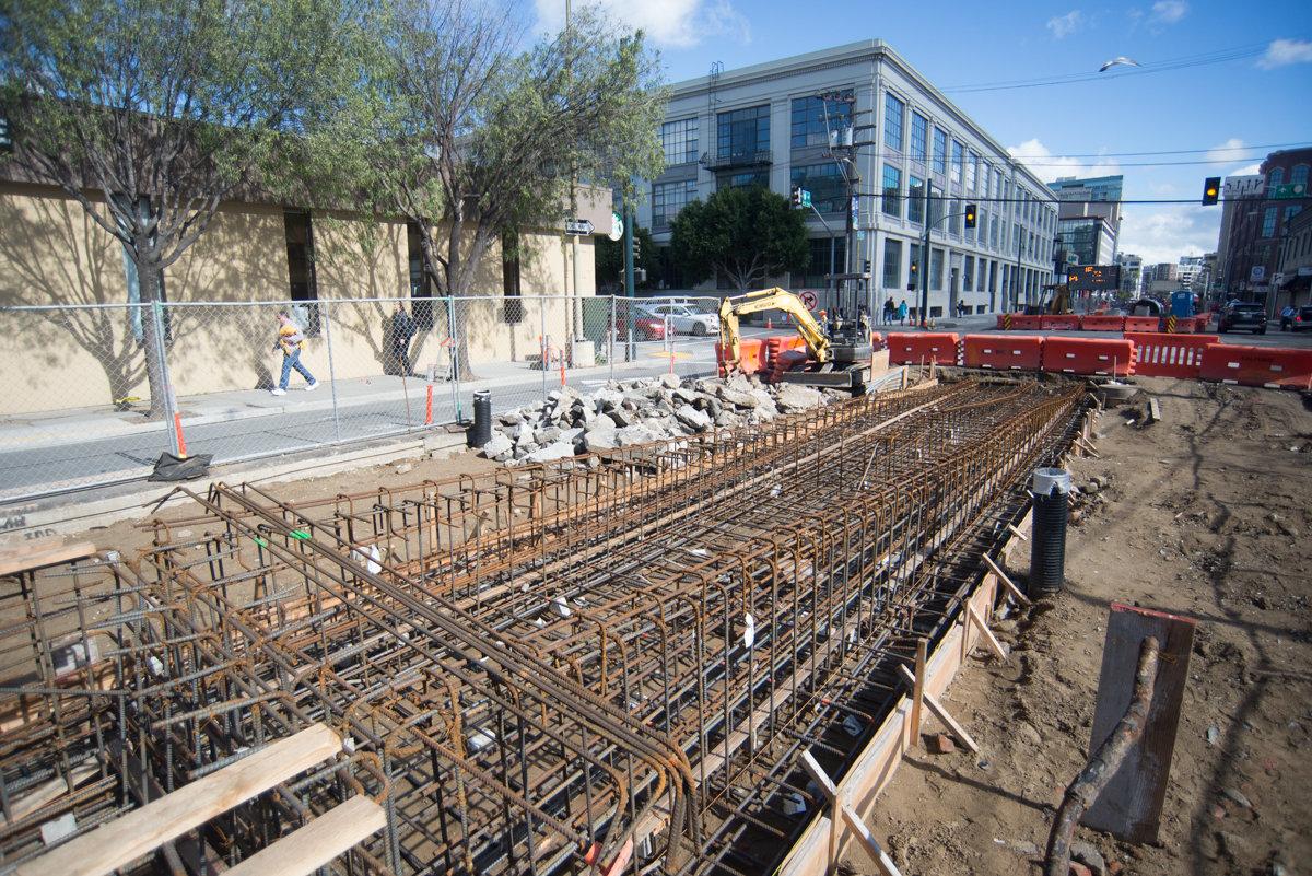 Construction site with rebar cages and excavation equipment at urban street intersection near buildings.