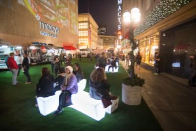 Nighttime urban plaza with crowds gathered around illuminated white seating cubes, storefronts and buildings visible in background.