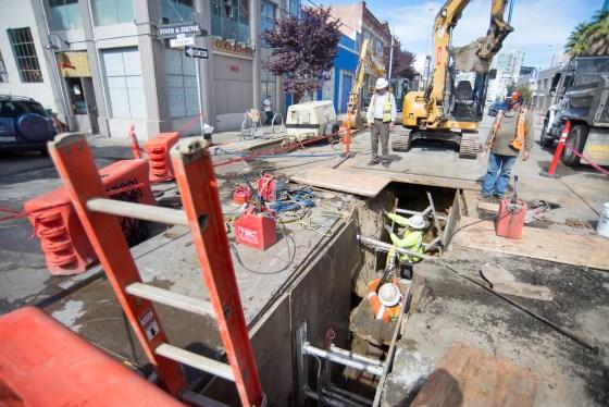 Construction crew performing underground utility work in street trench with excavator and safety equipment.