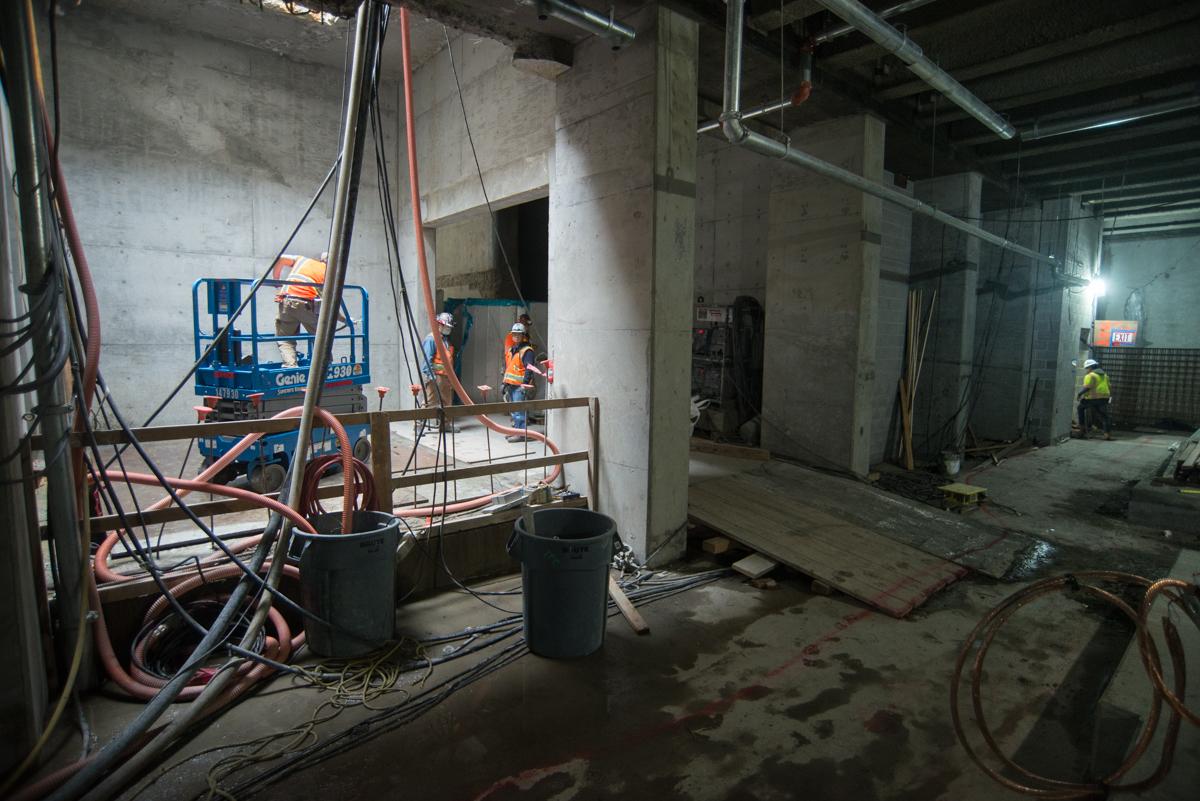 Construction crew operating scissor lift in underground transit station concourse with concrete pillars and utility equipment.