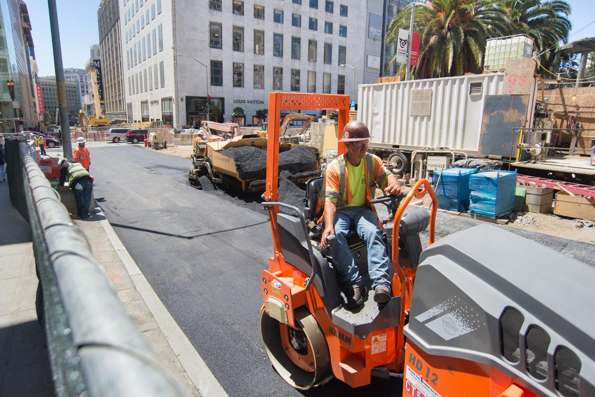 Construction worker operating orange steamroller on Stockton Street repaving project in downtown San Francisco.