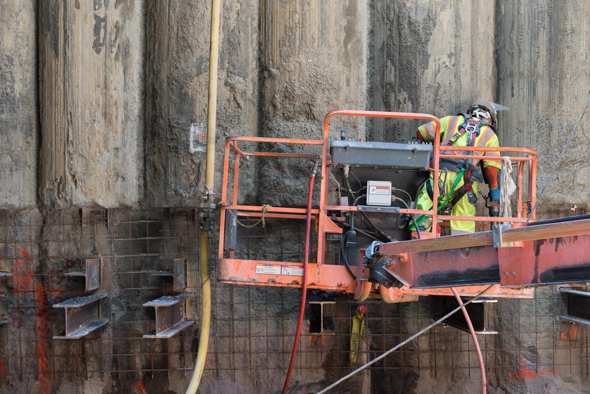 Worker on elevated platform preparing concrete wall for steel bracing installation at transit station excavation site.