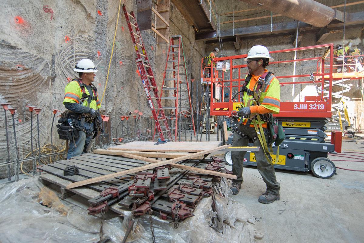 Construction workers in safety gear prepare slurry walls at a transit station headhouse, with scaffolding and equipment visible.