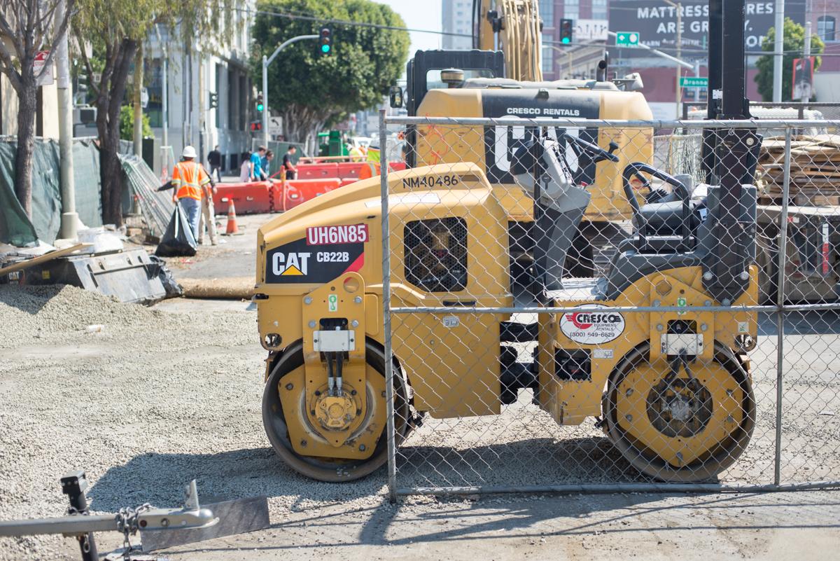 Yellow CAT steamroller and compactor equipment behind chain-link fence at urban construction site.