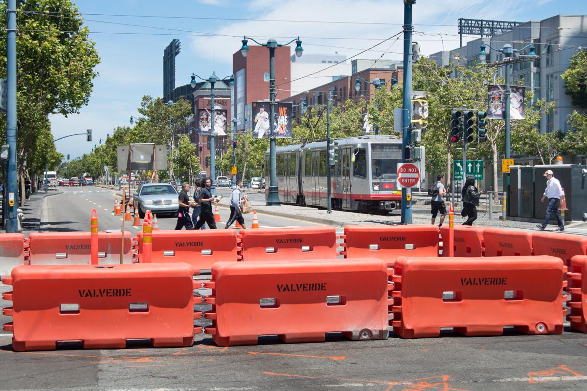 Red plastic barriers block a street intersection with a light rail vehicle and pedestrians visible in the background.