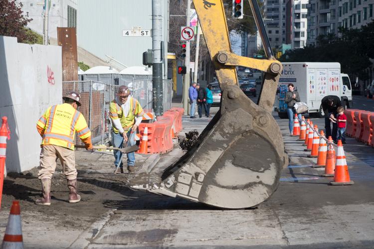 Construction workers excavate at 4th Street station using heavy equipment, with orange traffic cones and barriers lining the urban work site.