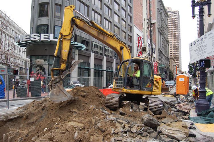 Yellow excavator with bucket digging into soil pile on urban street surrounded by buildings and construction barriers.