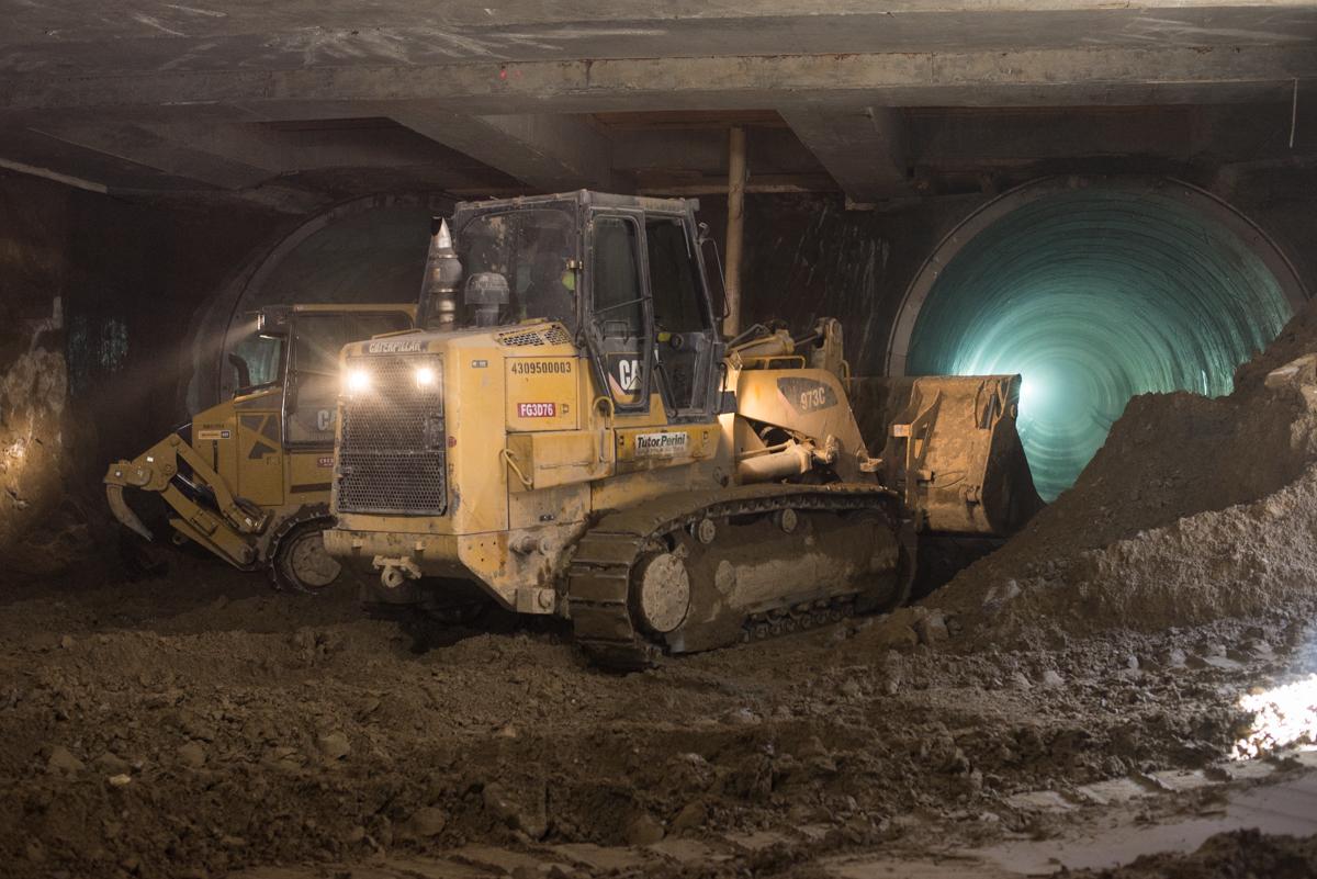 Bulldozer and excavation equipment working underground in tunnel during construction, with turquoise-lit pipe visible.