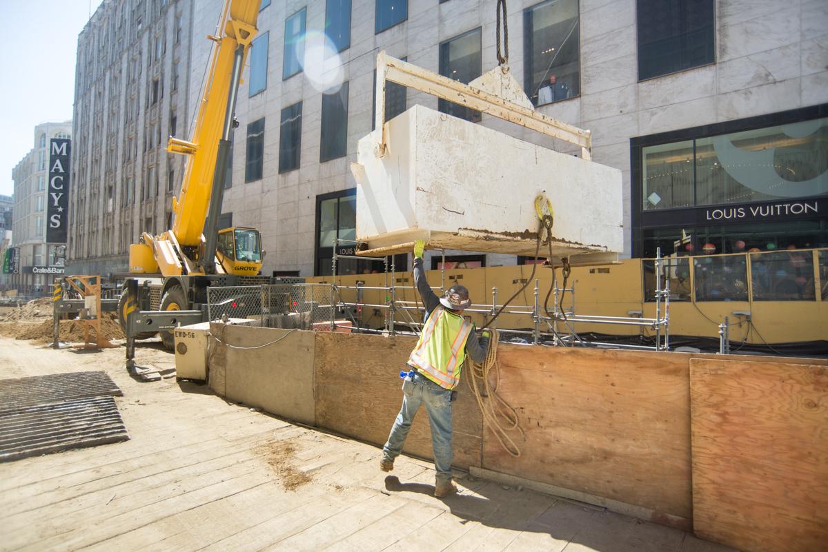 Construction worker guides steel bucket suspended by crane into underground station excavation on O'Farrell Street.
