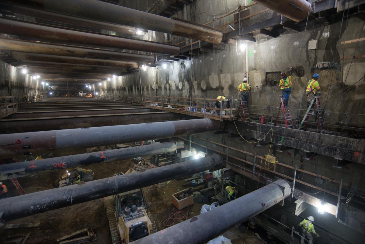Workers in hard hats and safety gear repair concrete walls inside an underground transit tunnel construction site.