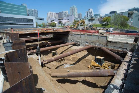 Construction excavation site showing exposed steel support beams and pipes with city skyline in background