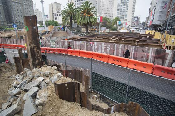 Construction site with sheet pile installation, orange barriers, and downtown buildings in background.
