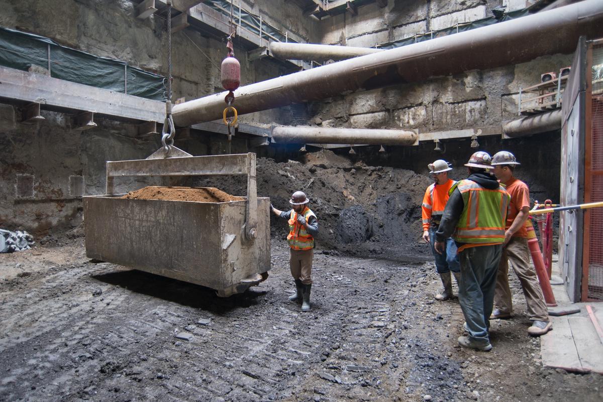 Construction workers in safety gear lower soil into a concrete bin at an underground transit construction site using a crane hook.