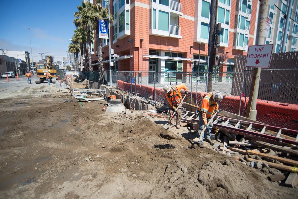Construction workers in orange vests perform utility work at a street site near a modern residential building.