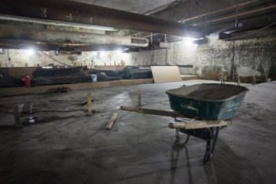 Industrial construction site with concrete floor, wheelbarrow, wooden supports, and exposed ceiling infrastructure.