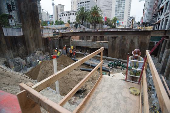 Construction excavation site on urban street with wooden framing, workers, and steel sheet piling along building foundation.