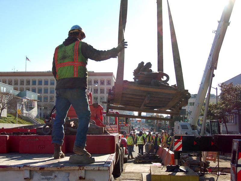 Worker in safety gear operating crane to lift steel beam during street construction in urban area.