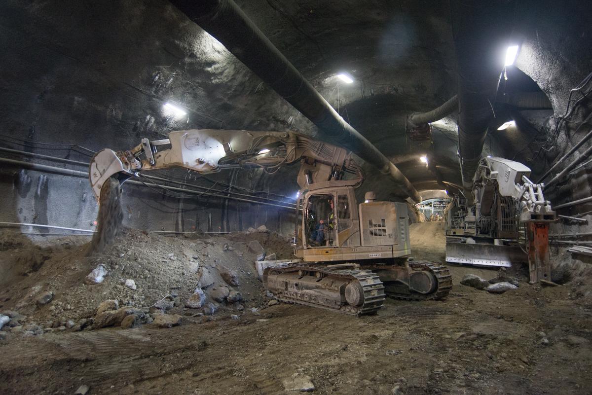 Excavator removing material inside underground station platform cavern with LED work lights and support structures visible.