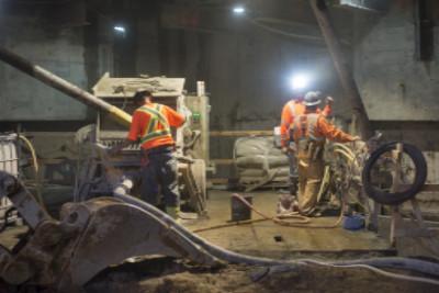 Workers in safety vests oversee shotcrete application in underground excavation at night.