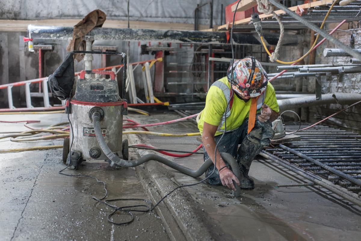 Worker in yellow safety vest kneeling on wet concrete floor operating pneumatic drill near construction equipment and steel reinforcement materials.