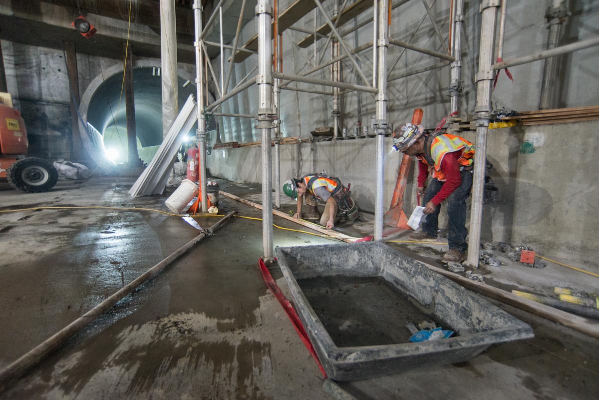 Workers in safety gear perform structural installation work on a station platform under scaffolding.