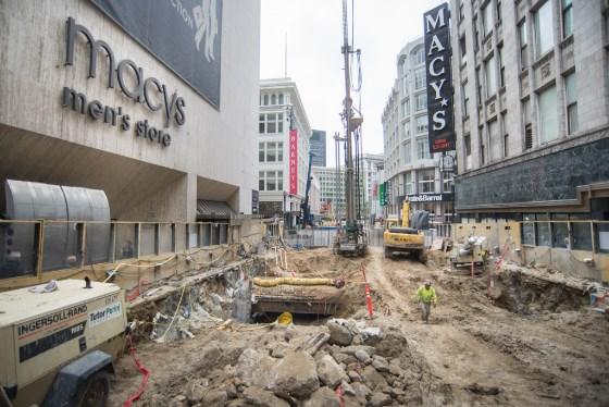 Construction excavation on downtown street with heavy equipment, closed storefronts, and surrounding buildings.