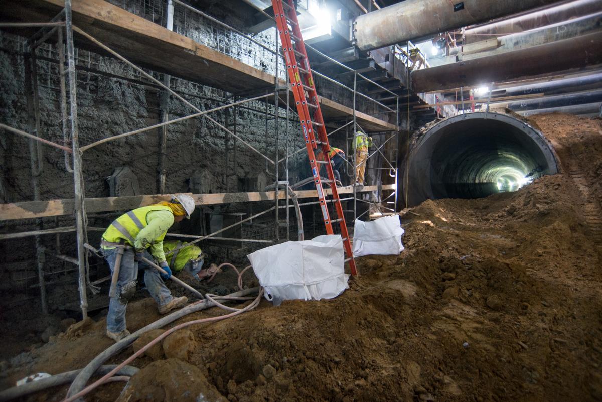 Worker in safety vest operating equipment inside underground transit station excavation with large tunnel pipe visible.