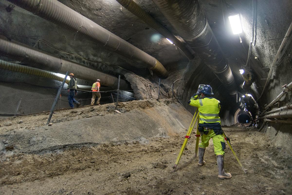 Workers in safety vests survey and excavate inside an underground transit tunnel with large pipes overhead and temporary support structures visible.