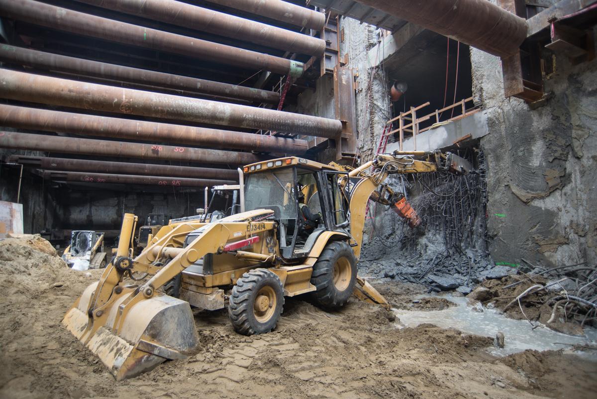 Yellow backhoe excavating debris at underground transit station construction site with steel pipes and concrete structure visible.