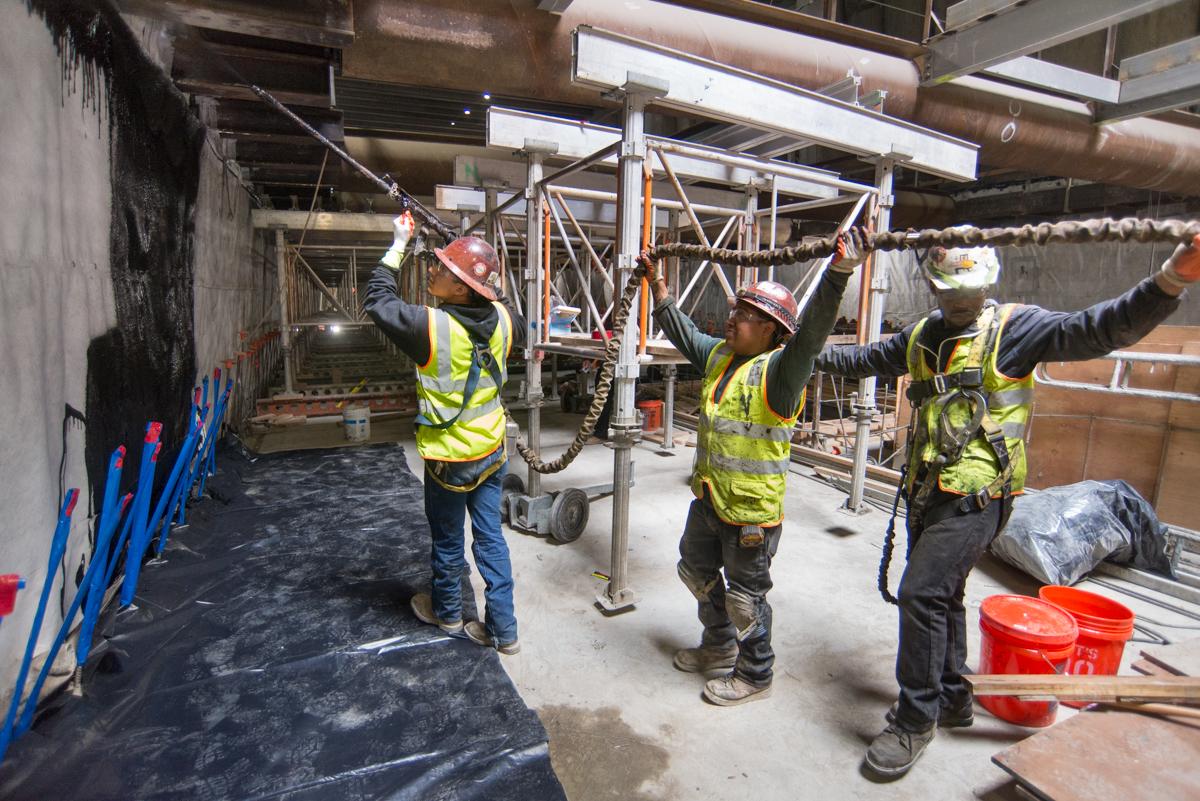 Construction workers in safety vests apply waterproofing material to interior wall during transit infrastructure repair.