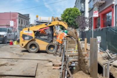 Yellow skid steer loader backfilling trench during sewer line construction on city street.