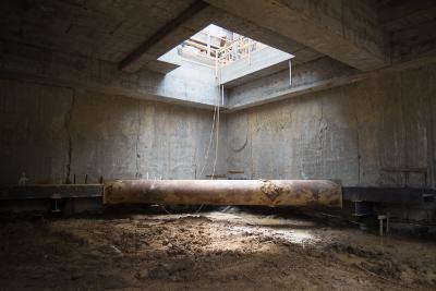 Concrete underground chamber with large pipe and roof opening allowing natural light, showing structural repair work in progress.