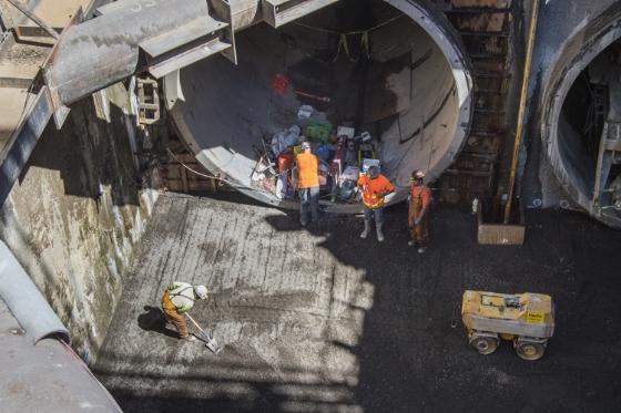 Workers in safety vests stand in a large circular tunnel shaft with concrete floor and yellow equipment visible.