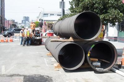 Large diameter sewer pipes stacked at a construction site with workers and equipment in the background.