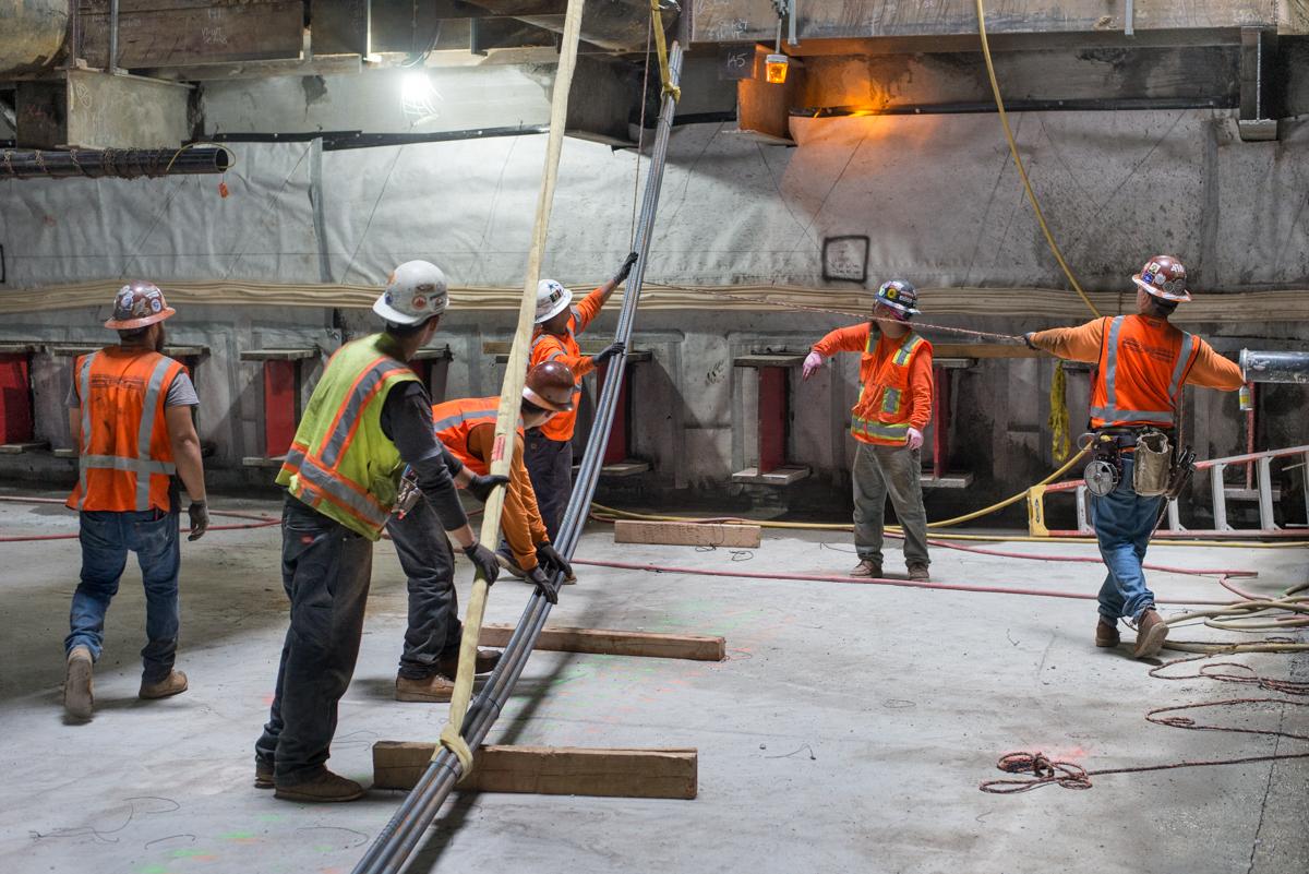 Construction workers in safety vests guide rebar being lowered into a transit station box at a work site.